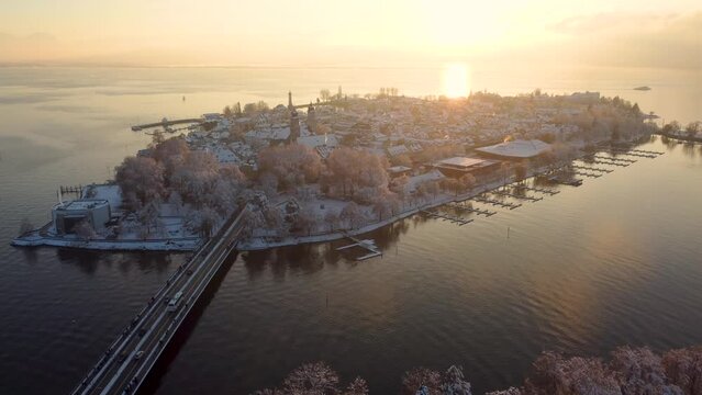 Lindau im Winter, warmer Sonnenuntergang am Bodensee über der Inselstadt Lindau, schneebedeckte Häuser der mittelalterlichen Stadt und Reif an den Bäumen. Klarer Horizont mit untergehender Sonne