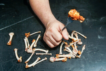 A man's hand among the gnawed chicken bones holds a fork on which part of a chicken wing is strung.