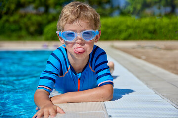 Funny little boy lies on the edge of a pool of blue water making grimaces. Funny kid with swimming...
