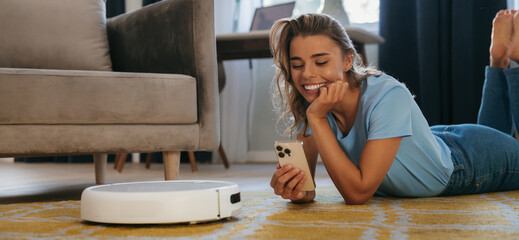 Smiling young woman lying down and using mobile phone to control smart vacuum cleaner robot