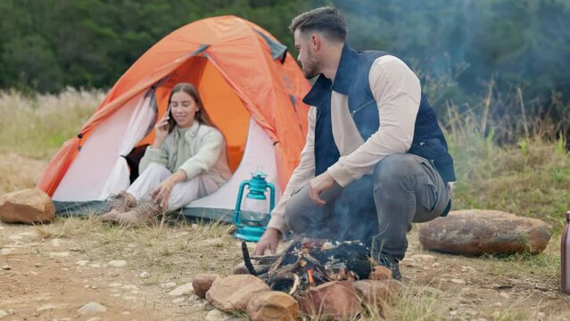 Couple, camping and outdoor with fire, talking and phone call to relax in woods, nature or forest on holiday. Man, woman and cellphone in tent for chat, cooking or vacation in bush, field or morning