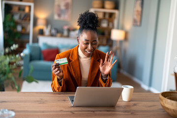 Smiling woman having video call at home holding credit card while sitting at desk with laptop and...