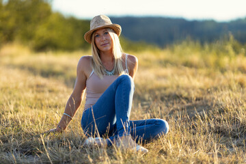 Young blonde woman wearing a straw hat, tank top and jeans sits leaning backwards on a harvested cornfield looking into the camera at sunset.