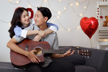 Happy Asian couple spending time together on sofa in living room, Guy playing acoustic guitar for...