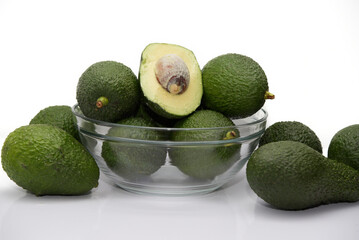 Close-up of a glass bowl full of green avocados and in the middle a halved avocado with the bone  in the air on a white background.