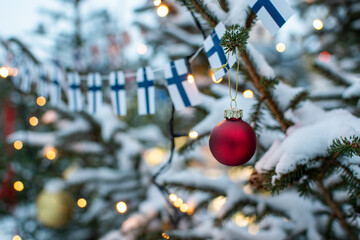 Red Christmas ball decor on a snow-covered Christmas tree with Christmas lights and Finnish flags