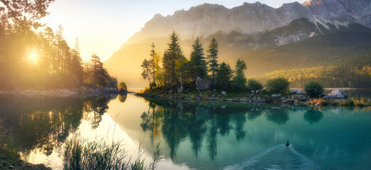 Idyllic lake at sunrise, a picturesque panorama with majestic mountains and the golden sunlight coming in behind the trees - obrazy, fototapety, plakaty