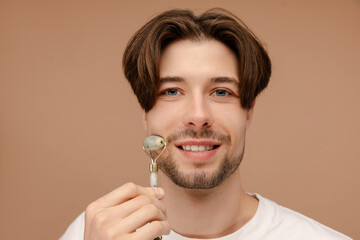 Portrait of happy young man massaging his face while holding jade roller. Isolated on beige...
