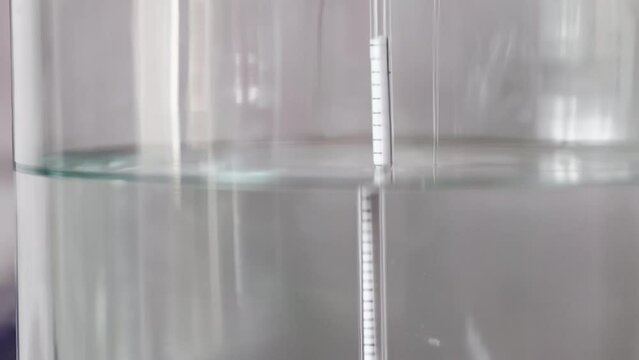 alcohol distilling process closeup - liquid distilled transparent liquid flowing in large glass jar with floating alcoholometer in it.