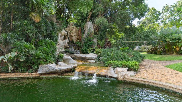 Location photos Waterfall garden in one of the tourist attractions. Daytime, clear weather, surrounded by nature, green leaves, large rocks, and stone paved walkways. Behind there is a helicopter.