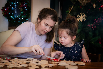 Mother and young daughter sit together and decorate Christmas cookies with icing