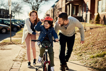 Family teaching son to ride bicycle on sidewalk