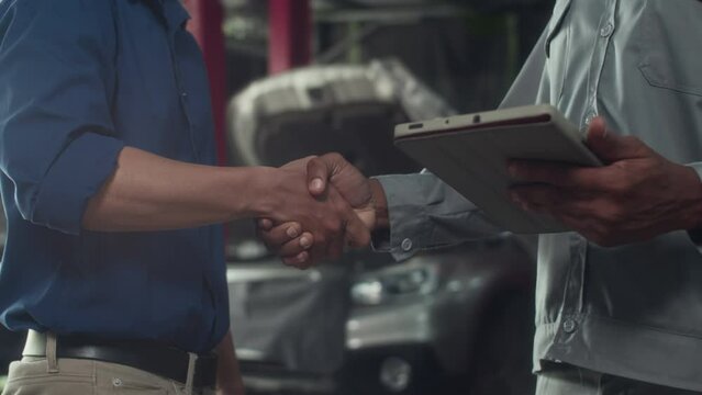 Handheld shot of specialist in gray uniform holding digital tablet shaking hand of client greeting at car repair service