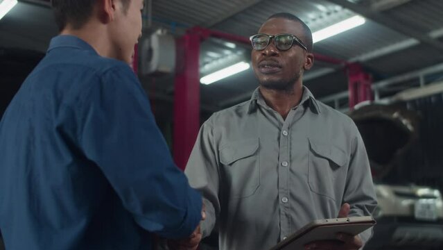 Low angle view of African American mechanic in gray uniform shaking hand of man coming to car serviceLow angle view of African American mechanic in gray uniform shaking hand of man coming to car servi