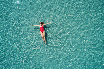 Obraz premium Top view of young pretty sexy girl in red swimsuit floating on water surface in crystal clear turquoise ocean. Happy island lifestyle.