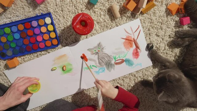 Process of painting family pet portrait with kid and cat lying near, top view. Four hands, art in process. Album on furry carpet, toy blocks around. Bright colors, nice simple paintings, HQ 4k footage