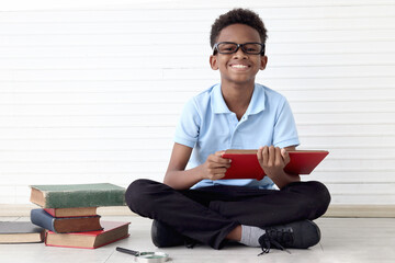 Happy smiling African boy with glasses reading book while sitting on floor in white wall room. Portrait of cute child with pile of books try to study, kid education, learning and studying.