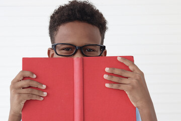 Happy African boy wearing glasses with opening red book on white wall room. Portrait of cute child covering his face with a book, kid education, reding, learning and studying concept.