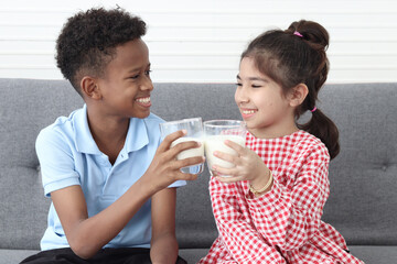 Happy smiling African boy and Asian girl drinking fresh milk together while sitting on sofa in...