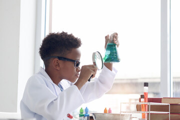 African boy in lab coat holds holding magnifying glass and chemical flask for doing science...