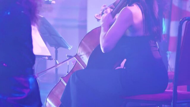 A female cellist playing in a concert hall. Close-up. Side view from behind. A cellist in a black evening dress. Light and smoke show