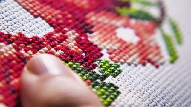 A close-up of a needle passing through the canvas and pulling a red thread behind it. The process of cross-stitch embroidery. Women's hobby. The concept of needlework