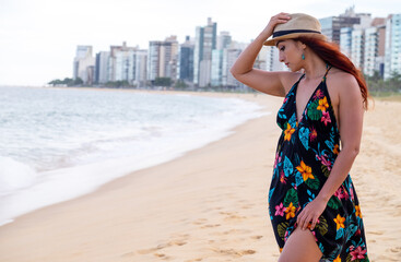 mujer con sobreso y vestido negro con flores en playa, arena y mar de fondo edificios de ciudad