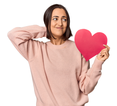 Young woman holding Valentine's heart touching back of head, thinking and making a choice.