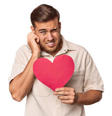 Young man holding a heart covering ears with hands.