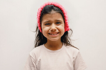 Cheerful Spanish girl child in a beige T-shirt and a pink headband on a white background, she shows her teeth, close-up, light, looking at the camera, 5 years old