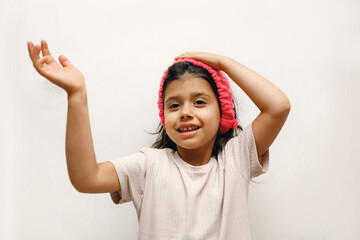 Cheerful Spanish girl child in a beige T-shirt and a pink headband on a white background, she shows...