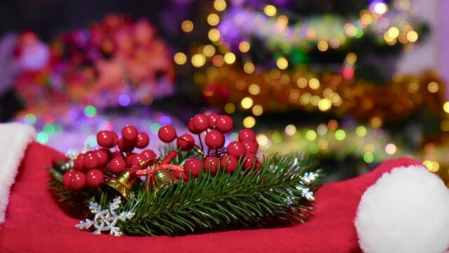 Close-up of Christmas tree branches with berries and bells lie on Santa Claus's hat on a Christmas background with flashing lights