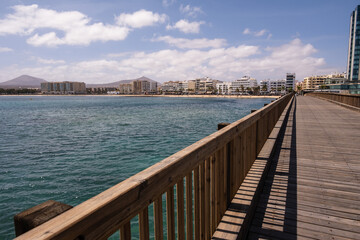 Fototapeta na wymiar View of the city of Arrecife from the Fermina islet, from a wooden bridge. Turquoise blue water. Sky with big white clouds. Seascape. Lanzarote, Canary Islands, Spain.
