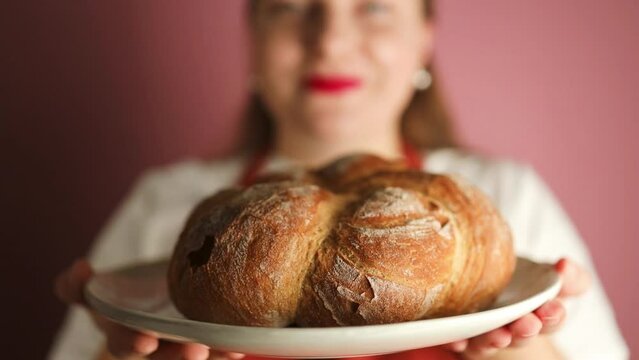 Portrait of young happy female baker in apron holding a plate with fresh bread and smiling at camera isolated over pink background. Healthy organic food. High quality FullHD footage