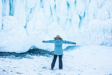 Female in warm thermal clothing stands with her back and open arms against big ice on the rocks 