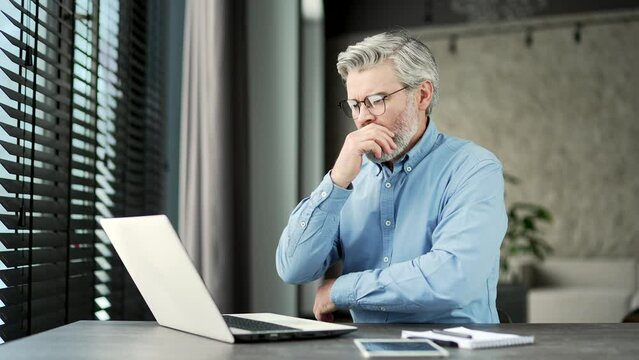 Frustrated stressed mature gray haired bearded businessman reads bad news on a laptop while sitting at workplace in business office. Disappointed entrepreneur is upset reviewing negative information