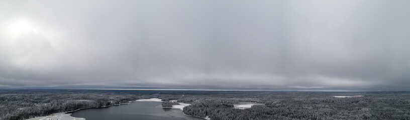 Aireal panoramic view of extensive grey clouds above a snowy forest and a partially frozen lake. The winter scene has a cold feeling.
