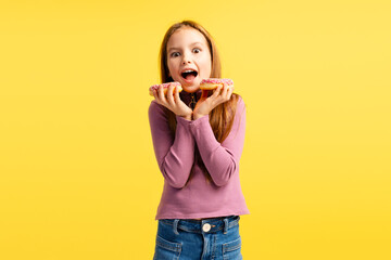 Portrait of attractive excited little girl wearing casual clothes, eating tasty pink donut, standing isolated on yellow background. Concept of food, dessert - Powered by Adobe