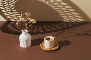 Creative composition of white vase with dry flowers and cup of coffee on brown and pastel beige...