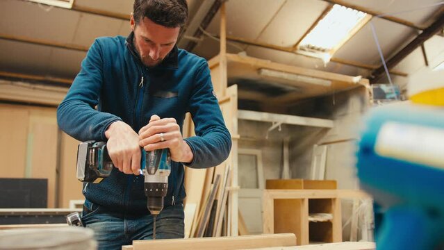 Male carpenter at workbench drilling hole in piece of wood with cordless electric drill - shot in slow motion
