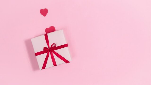 4k Red hearts, symbols of love, fly out of gift box tied with a red ribbon. Greeting card for valentine's day. Pink background. Copy space. Flat lay. Stop motion animation.