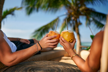 Couple enjoys fresh coconut water on tropical beach, seated under palm tree. Man and woman toast coconuts, sipping drinks, ocean view. Romantic getaway, leisure moment, chill vacation vibe. - obrazy, fototapety, plakaty