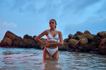 Attractive woman enjoys nighttime swim in ocean under crimson illumination. Fashionable lady in swimwear poses during an evening ocean dip.