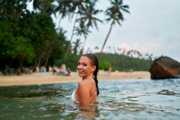Woman enjoys swimming in ocean. Female with waterproof makeup smiles in tropical sea. Fashionable swimwear on vacation. Joyful traveler in natural environment. Lady explores beach lifestyle.