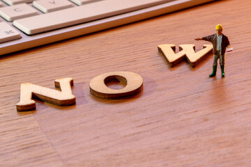 NOW word or concept made by wooden letters on wooden background with white keyboard in the background and miniature workers figurines adding W constructing NOW