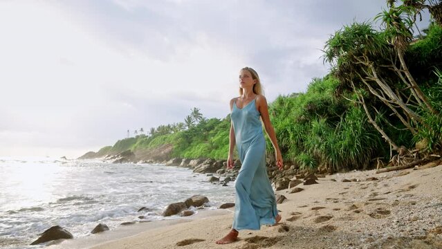 Bronzed woman in elegant blue gown strolls sandy beach. Fashion model showcases summer dress, seaside ambiance. Luxury travel lifestyle, tropical holiday. Fashion shoot, perfume ads potential.