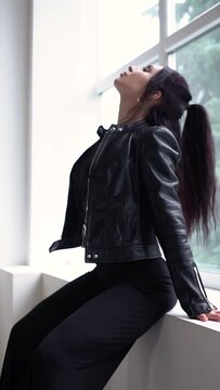 A hot sexy brunette girl in a black dress and a black leather jacket sits on a window in a white studio and looks at the camera