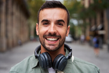 Portrait of a young handsome man smiling and looking at camera. Happy male wearing headphones in street on the city. Close up of a cheerful millennial caucasian guy outdoors. - obrazy, fototapety, plakaty