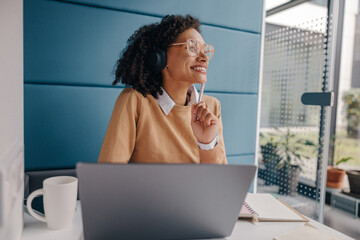 Smiling female freelancer in headphones listen music and looks away while sitting in coworking 