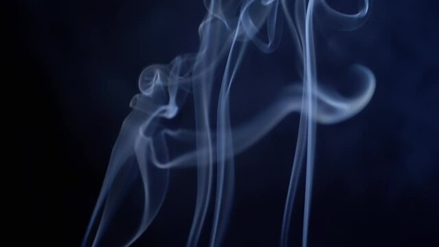 Thin Curls of Blue Smoke Rise Up, Fill Empty Space on a Black Background. Incense sticks. Abstract shapes. Texture. Beautiful clouds of smoke swirl. Floating fog, flowing smoke. Blurred motion. Light.